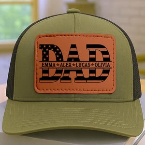 May include: A green and black trucker hat with a brown leather patch. The patch features the word "DAD" in a bold font with an American flag design, and the names "EMMA ALEX LUCAS OLIVIA" below. The hat is sitting on a stack of books.