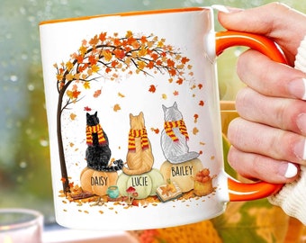 Puede incluir: Taza de cerámica blanca con asa e interior naranja. La taza presenta una ilustración con temática otoñal de tres gatos sentados sobre calabazas bajo un árbol con hojas cayendo. Los gatos se llaman Daisy, Lucie y Bailey.