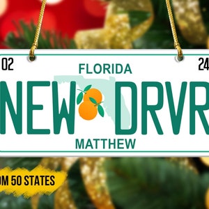 May include: A white and green Florida license plate ornament with the text "NEW DRVR" and the name "MATTHEW". The ornament features a stylized outline of the state of Florida with two orange citrus fruits.