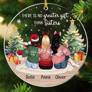 Puede incluir: Un adorno navide&ntilde;o redondo con una escena de invierno con tres mujeres con su&eacute;teres y gorros de Pap&aacute; Noel. El texto "There is no greater gift than sisters" est&aacute; impreso en el adorno. Los nombres "Bella, Anna, Oliver" est&aacute;n impresos en la parte inferior.