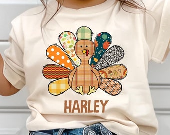 May include: Cream-colored t-shirt featuring a colorful turkey design with various patterns and textures. The turkey has a plaid body and patterned feathers in orange, green, and brown. The name "HARLEY" is printed below the turkey in brown.
