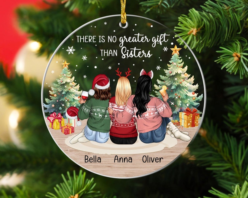 Puede incluir: Un adorno navide&ntilde;o redondo con una escena de invierno con tres mujeres con su&eacute;teres y gorros de Pap&aacute; Noel. El texto "There is no greater gift than sisters" est&aacute; impreso en el adorno. Los nombres "Bella, Anna, Oliver" est&aacute;n impresos en la parte inferior.