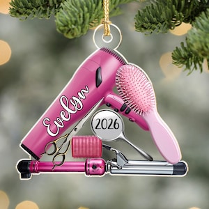 Puede incluir: Un adorno colgante de forma triangular plana que presenta un secador de pelo rosa con el nombre Evelyn impreso en letra blanca, un cepillo para el cabello rosa, un rulo rosa, un espejo de mano plateado con el a&ntilde;o 2026 y un rizador de pelo.