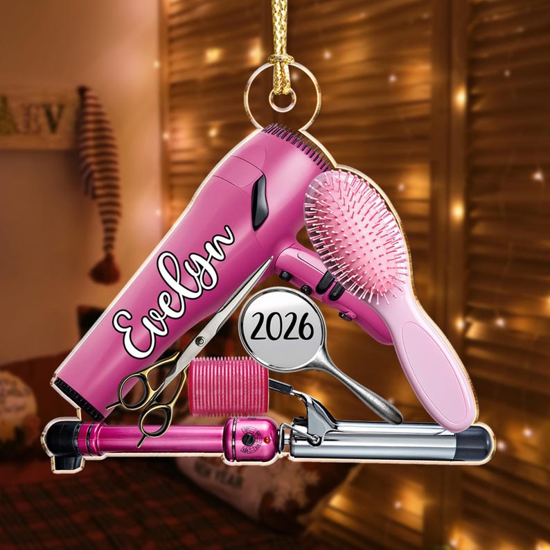 Puede incluir: Un adorno colgante que muestra un secador de pelo rosa con el nombre Evelyn impreso en letra blanca, un cepillo rosa, un espejo de mano plateado con el a&ntilde;o 2026, un rulo rosa y unas tijeras de peluquer&iacute;a plateadas, sujeto por un cord&oacute;n dorado.