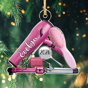 Puede incluir: Un adorno colgante que presenta un secador de pelo rosa con el nombre Evelyn impreso, un cepillo de pelo rosa, un rulo rosa, unas tijeras doradas y una etiqueta circular que muestra el a&ntilde;o 2026, todo unido a un cord&oacute;n dorado para colgar.