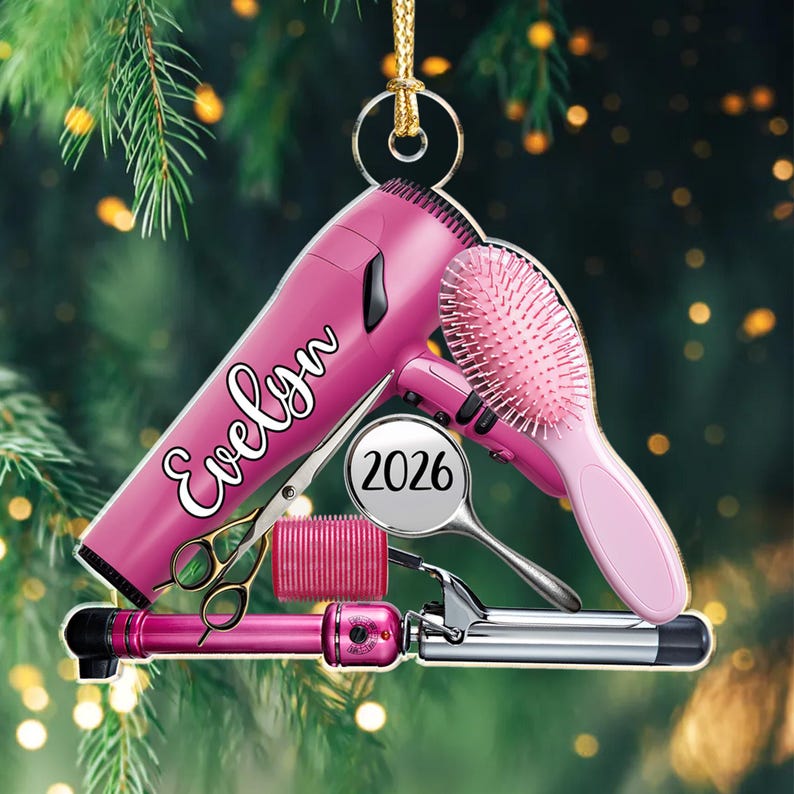 Puede incluir: Un adorno colgante que presenta un secador de pelo rosa con el nombre Evelyn impreso, un cepillo de pelo rosa, un rulo rosa, unas tijeras doradas y una etiqueta circular que muestra el a&ntilde;o 2026, todo unido a un cord&oacute;n dorado para colgar.