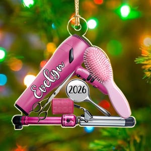 Puede incluir: Un adorno de secador de pelo rosa con el nombre Evelyn en letra blanca, un cepillo rosa, un rulo rosa, unas tijeras de peluquer&iacute;a plateadas y una tenacilla plateada con una etiqueta circular que muestra el a&ntilde;o 2026, colgado de un cord&oacute;n dorado.