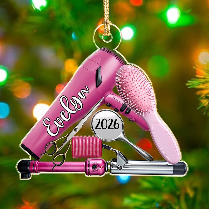 Puede incluir: Un adorno de secador de pelo rosa con el nombre Evelyn en letra blanca, un cepillo rosa, un rulo rosa, unas tijeras de peluquer&iacute;a plateadas y una tenacilla plateada con una etiqueta circular que muestra el a&ntilde;o 2026, colgado de un cord&oacute;n dorado.