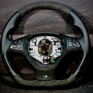 May include: Black steering wheel with perforated leather grips and suede-like sections. The wheel features integrated controls and a central hub with exposed wiring. The M logo is visible. The wheel is designed for a vehicle.