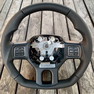 Puede incluir: Un volante negro con un agarre de cuero perforado. El volante tiene botones de control integrados en los lados izquierdo y derecho, con un cubo central con componentes mecánicos expuestos.