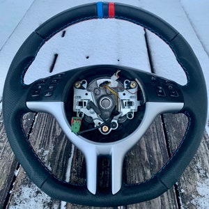 May include: Black leather steering wheel with red, blue, and white accents. The wheel has a perforated grip and red and blue stitching. The center of the wheel is silver and has various buttons and controls.