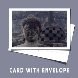 Op de afbeelding: Een ansichtkaart met een verweerde stenen Boeddha-standbeeld met een serene uitdrukking, tegen een achtergrond van een stenen structuur. De kaart is wit en bevat de tekst "CARD WITH ENVELOPE" in een schreefloos lettertype.