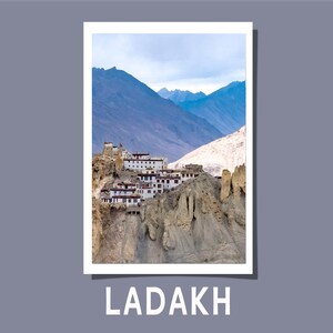 May include: A photograph of a mountain landscape featuring a cluster of white buildings with red accents perched on a rocky cliff. The background showcases towering blue mountains under a cloudy sky. The word "LADAKH" is printed in white at the bottom.