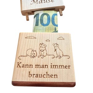 Könnte beinhalten: Ein Geldgeschenkhalter aus hellem Holz, bestehend aus einem Oberteil mit der Gravur "Mäuse" und einem Unterteil mit drei Mäusen und dem Text "Kann man immer brauchen," wobei ein 100-Euro-Schein zwischen den beiden Holzteilen eingesteckt ist.