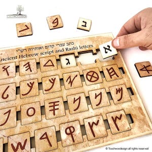 May include: A wooden puzzle with beige tiles featuring Hebrew letters in brown. The tiles are arranged in a grid pattern. The text "כתב עברי קדום ואותיות רש"י" is written in Hebrew at the top of the puzzle. The text "Ancient Hebrew script and Rashi letters" is written in English below the Hebrew text.