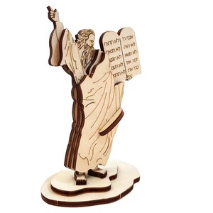 May include: A wooden sculpture depicting a robed figure holding tablets inscribed with Hebrew text. The figure has a long beard and is made from light brown wood. It stands on a wooden base.