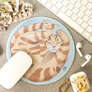 Op de afbeelding: Een ronde muismat met een lichtblauwe achtergrond en een cirkelvormige illustratie van een rosse cyperse kat die zich tot een bal heeft opgerold met de pootjes omhoog. De kat heeft gesloten ogen en een vredige uitdrukking in het gezicht.