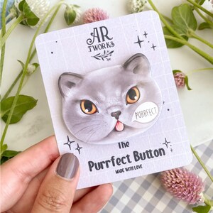 Pode incluir: Um botão de gato cinza com um balão de fala rosa que diz "Purrfect". O botão está em um cartão branco com o texto "The Purrfect Button" e "AR TWORKS" em texto preto.