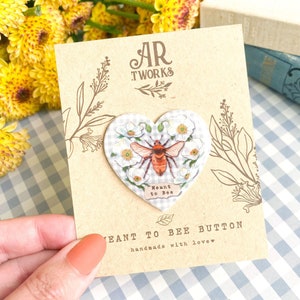 Pode incluir: Um botão em forma de coração com uma ilustração de abelha e o texto "Meant to Bee". O botão está sobre um cartão de papel kraft com o texto "Meant to Bee Button". O fundo apresenta flores brancas, flores amarelas e um padrão xadrez azul e branco.