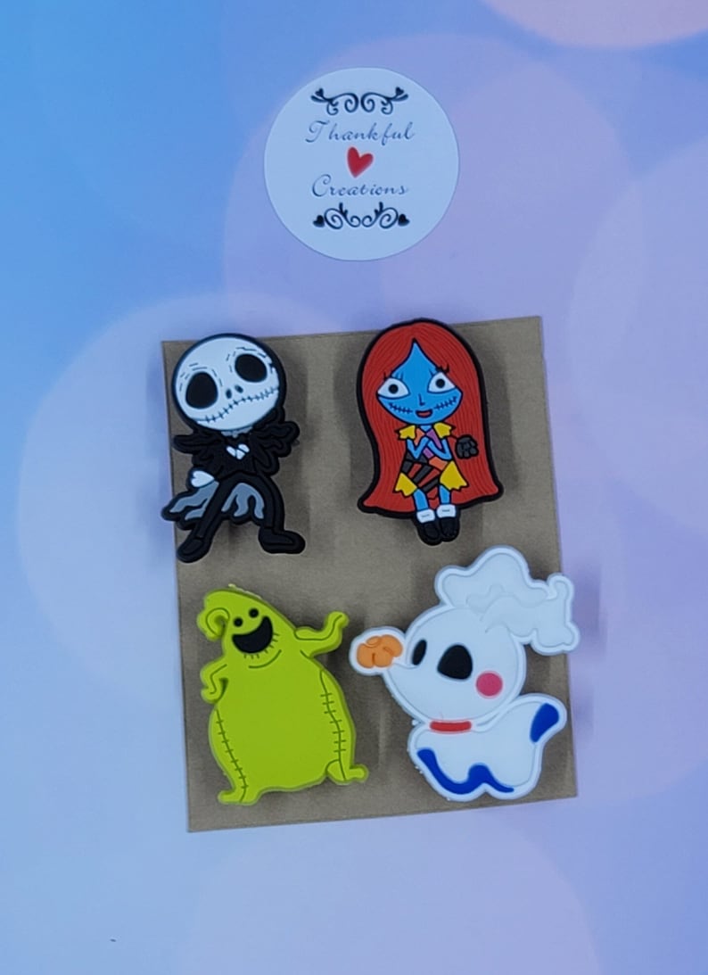 May include: A set of four colorful rubber shoe charms featuring characters from The Nightmare Before Christmas, including a skeleton, a red-haired figure, a green sack-like creature, and a white ghost dog, with a Thankful Creations logo sticker.