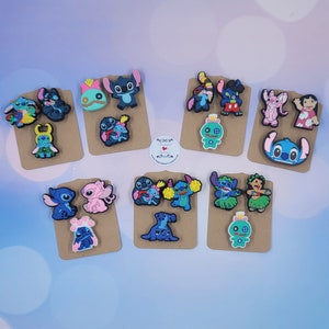 May include: A collection of colorful rubber shoe charms featuring various cartoon characters, including blue and pink creatures, displayed on brown cardboard backing cards. The charms depict characters in different poses, outfits, and expressions.