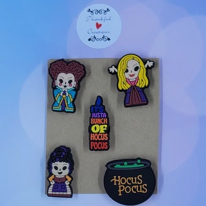 May include: A set of five colorful rubber shoe charms featuring three cartoon characters, a black candle with the text IT'S JUSTA BUNCH OF HOCUS POCUS, and a black cauldron with green liquid and the text HOCUS POCUS, all arranged on a brown card.