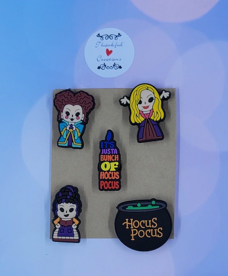 May include: A set of five colorful rubber shoe charms featuring three cartoon characters, a black candle with the text IT'S JUSTA BUNCH OF HOCUS POCUS, and a black cauldron with green liquid and the text HOCUS POCUS, all arranged on a brown card.