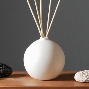 Könnte beinhalten: Weiße Keramik-Diffuserflasche mit Holzstäbchen, die auf einer Holzoberfläche mit zwei grauen Bimssteinen steht.