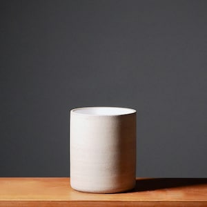 Peut inclure: Un vase cylindrique en céramique blanche avec une surface lisse et légèrement texturée. Le vase est posé sur une surface en bois.