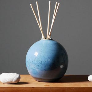Peut inclure: Un diffuseur en céramique bleu avec des bâtonnets en bois, présenté avec des pierres décoratives. Le vase sphérique a un glaçage tacheté. Les bâtonnets sont brun clair et s'étendent vers le haut. Les pierres sont grises et blanches.
