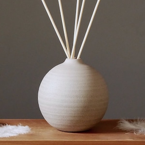 Könnte beinhalten: Eine runde, hellgraue Diffuserflasche aus Keramik mit einem Satz Holzdiffuserstäbchen. Die Flasche steht auf einer Holzoberfläche mit weißen Federn.