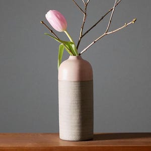 Op de afbeelding: Een licht roze en grijze keramische vaas met een enkele roze tulp en takken.