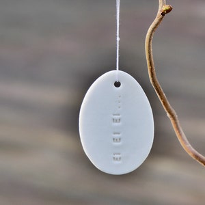 Pode incluir: Ornamento de ovo de cerâmica branca com a palavra "Acreditar" em relevo, pendurado em um cordão branco em um galho marrom.