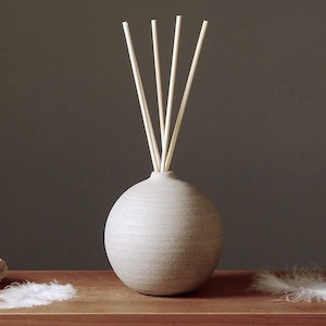 May include: A gray ceramic diffuser bottle with a round shape and a set of wooden sticks. The bottle is sitting on a wooden surface with a white feather on either side.
