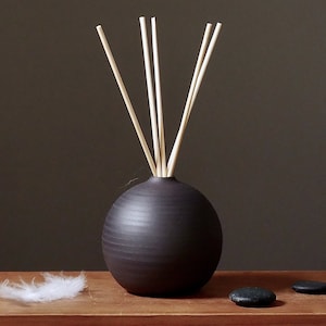 May include: A dark brown, spherical ceramic diffuser with several light-coloured reeds. The diffuser sits on a wooden surface, with a white feather and two black stones nearby. The background is a solid dark brown.