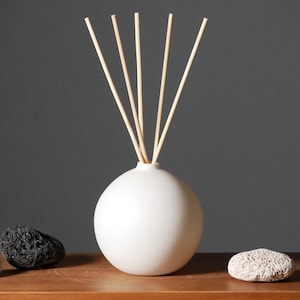 Könnte beinhalten: Eine weiße Keramik-Diffuserflasche mit runder Form und Holzstäbchen, die aus der Oberseite herausragen. Die Flasche steht auf einer Holzoberfläche mit zwei kleinen grauen Steinen.