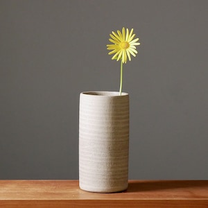 Könnte beinhalten: Eine einzelne gelbe Gänseblümchen-Blume in einer grauen Keramikvase mit strukturierter Oberfläche. Die Vase steht auf einem Holztisch.
