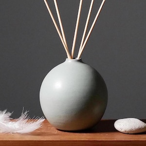 Könnte beinhalten: Eine hellgraue Keramik-Diffuserflasche mit Holzstäbchen, die aus der Oberseite herausragen. Die Flasche ist rund und hat eine glatte Oberfläche. Eine weiße Feder und ein grauer Stein befinden sich im Vordergrund.