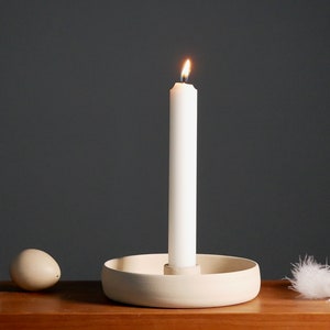May include: A white candle burning in a white ceramic candle holder with a white feather on a wooden surface.