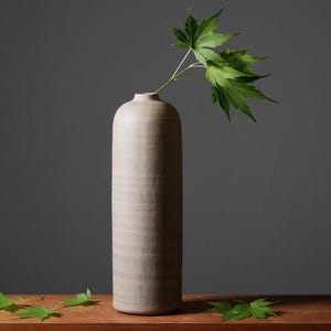 Op de afbeelding: Een hoge, cilindervormige beige vaas met een textuur houdt een takje groene bladeren vast. De vaas staat op een houten oppervlak, met een donkergrijze achtergrond. Verschillende groene bladeren liggen op het houten oppervlak.