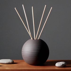 Könnte beinhalten: Eine dunkelbraune, kugelförmige Keramikvase mit strukturierter Oberfläche, die mehrere hellfarbene Schilfrohr-Diffusorstäbchen enthält. Drei glatte, graue Steine sind auf einer Holzoberfläche vor der Vase angeordnet. Der Hintergrund ist einfarbig dunkelgrau.