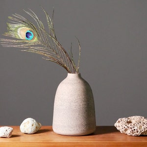 Könnte beinhalten: Eine graue Keramikvase mit strukturierter Oberfläche hält eine einzelne Pfauenfeder mit einem blauen und grünen Auge. Die Vase steht auf einem Holztisch mit drei kleinen weißen Steinen.