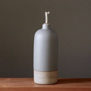 Könnte beinhalten: Eine hohe, graue Keramikölflasche mit einem silbernen Ausgießer. Die Flasche hat einen abgerundeten oberen Teil und einen hellbeigen Sockel. Das Design ist minimalistisch und modern, passend für die Kücheneinrichtung.
