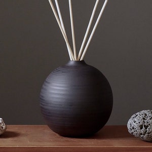 Könnte beinhalten: Eine schwarze Keramik-Diffuserflasche mit runder Form und einer strukturierten Oberfläche. Die Flasche hat einen Satz weißer Diffusorsticks, die oben eingesetzt werden.