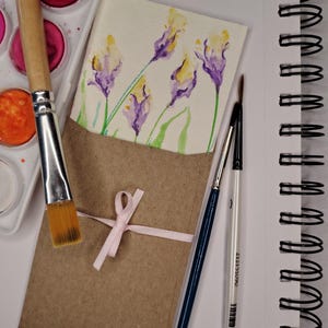 May include: A watercolor painting of purple and yellow flowers on a rectangular card tucked into a brown paper envelope tied with a light pink ribbon. A paint palette with various colors, two paintbrushes, and a spiral-bound sketchbook are present.