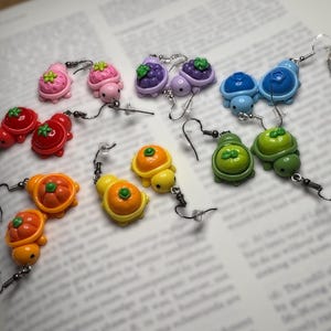 Peut inclure: Une collection de boucles d'oreilles fantaisie colorées représentant des fruits et légumes miniatures, notamment des fraises, des myrtilles, des tomates, des oranges et des tortues. Les boucles d'oreilles sont en matériau brillant.