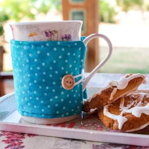 Puede incluir: Una taza de cerámica blanca con un diseño floral y un calientavasos de tela azul con lunares. La taza está sobre una bandeja blanca con dos galletas glaseadas.