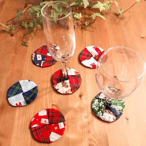 May include: Six colourful fabric coasters with a Christmas theme. The coasters feature a variety of patterns, including plaid, polka dots, and snowflakes. Two clear glass glasses are sitting on top of the coasters.