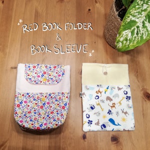 Puede incluir: Dos fundas de libro de tela. Una es una carpeta roja para libros con un patrón floral y un bolsillo rosa. La otra es una funda de libro con fondo blanco y un estampado de animales de colores. El texto "RED BOOK FOLDER & BOOK SLEEVE" está escrito encima de las fundas.