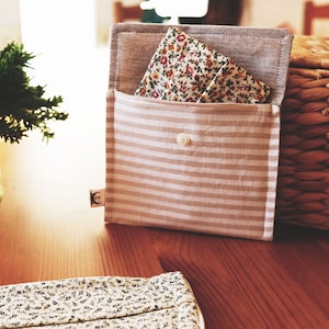 Puede incluir: Una bolsa de tela a rayas beige y blancas con una tela estampada floral en el interior. La bolsa se cierra con un botón blanco. Una pequeña planta verde en una maceta estampada y una cesta de mimbre están en el fondo.